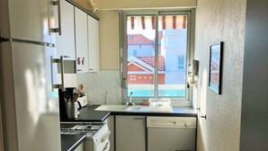 A compact kitchen with a refrigerator, stove, sink, and dishwasher. A window provides a view of buildings outside.