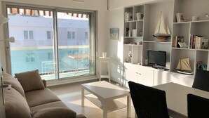 A modern living room with a sofa, a dining table, and chairs. There is a bookshelf with various items and a television. A large window offers a view of buildings outside.