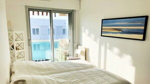 A bedroom with a bed, a nightstand, a lamp, a window with a view of buildings, and a framed picture on the wall.