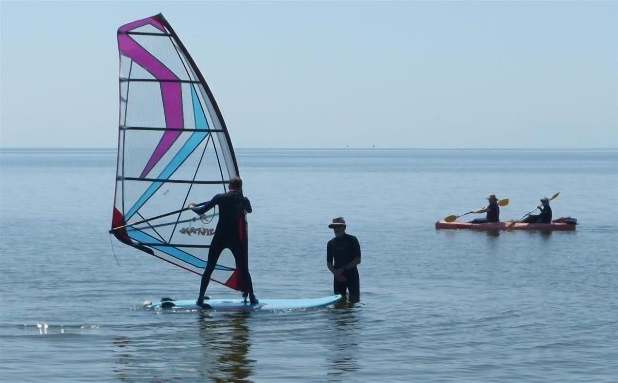 A person windsurfing on a calm sea with a colorful sail, another person paddling a kayak, and a third person standing in the water.