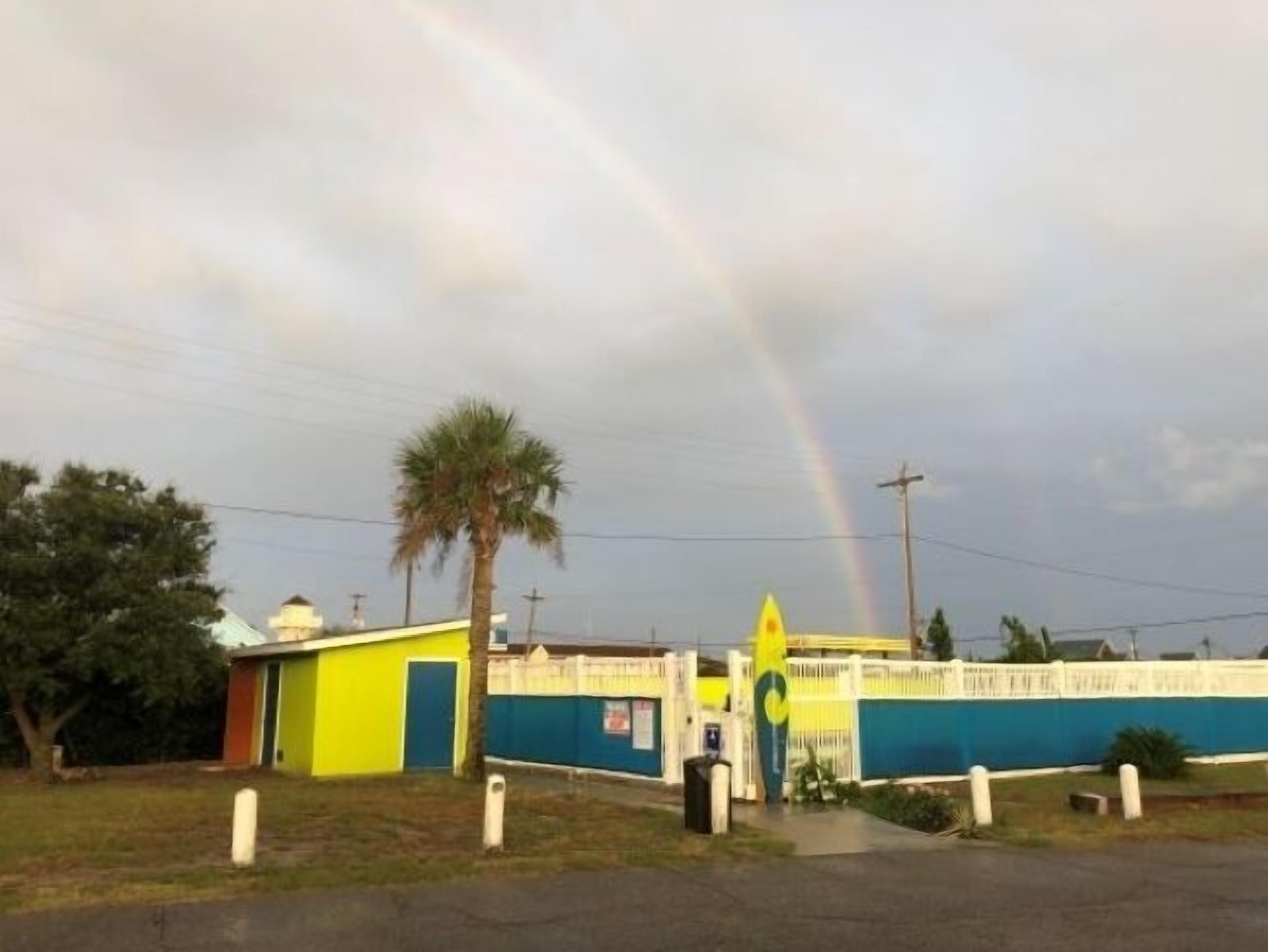 A building with a yellow and blue exterior, a palm tree, and a rainbow in the sky.