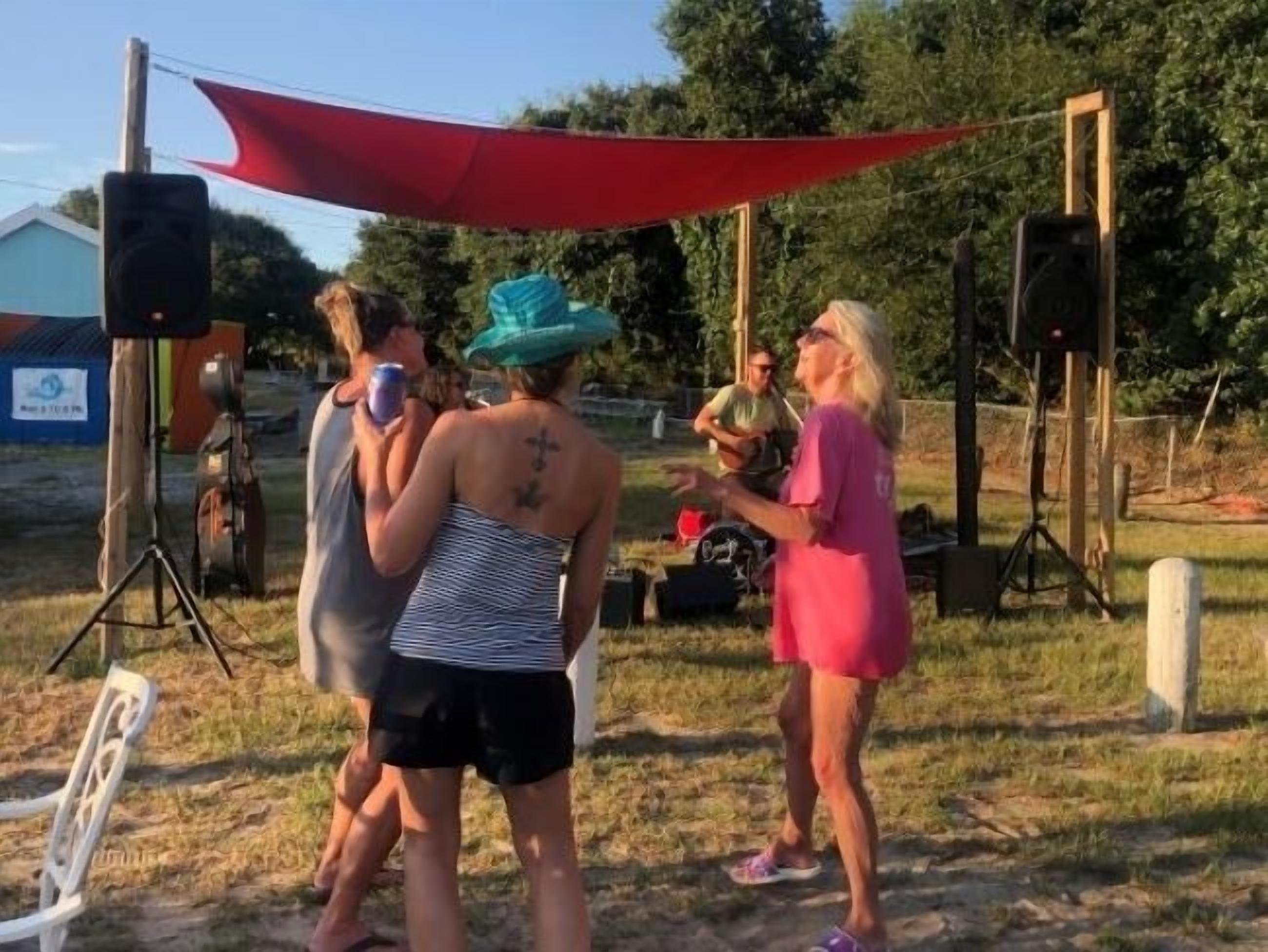 A group of people outdoors, possibly at a social event or gathering, with a red canopy overhead and various equipment like speakers and a tripod visible.