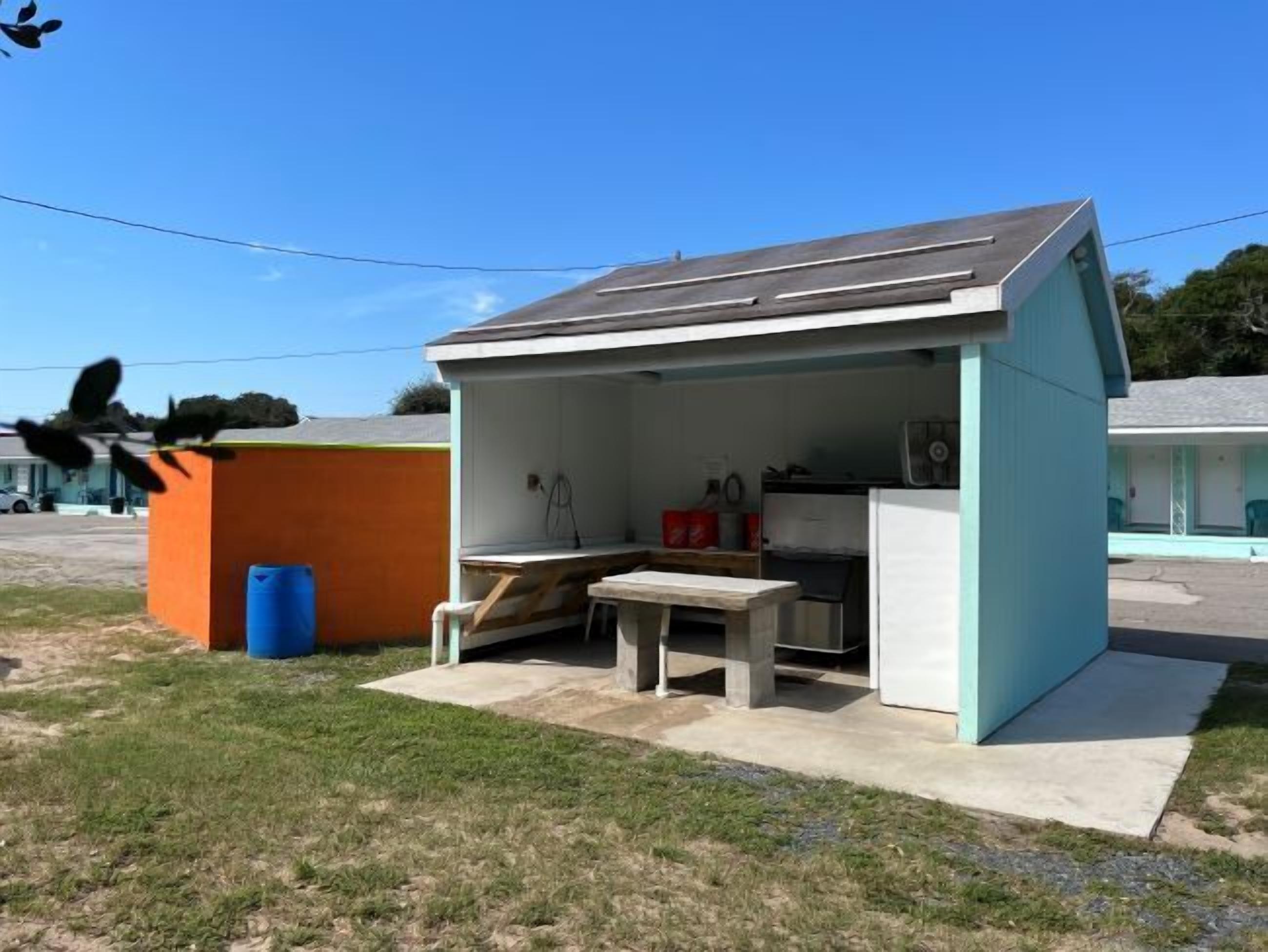 A small, single-story building with a sloped roof, a concrete patio, and a simple outdoor seating area.