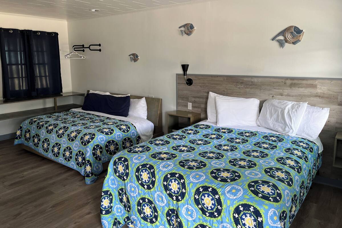 A hotel room with two beds, a wooden headboard, a nightstand, a wall-mounted lamp, and a window with curtains.