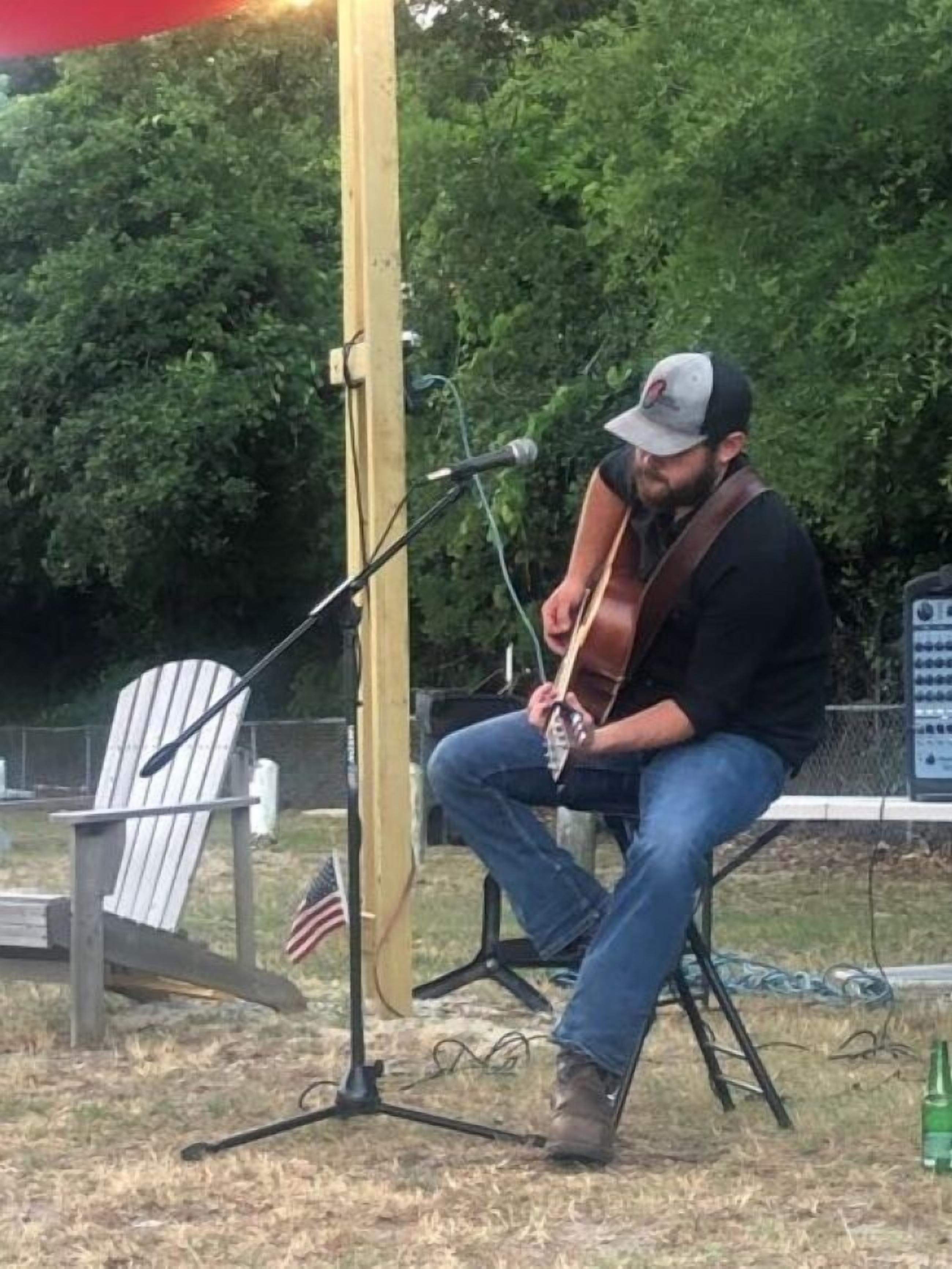 Una persona tocando la guitarra en una silla al aire libre, con un soporte de micrófono al frente, una bandera estadounidense y un altavoz portátil al fondo.