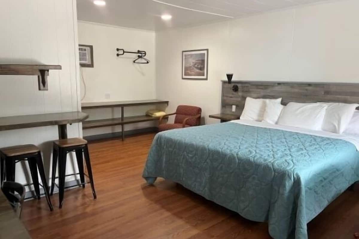 A hotel room with a bed, two bar stools, a chair, a wooden headboard, a wall-mounted coat rack, and a framed picture.