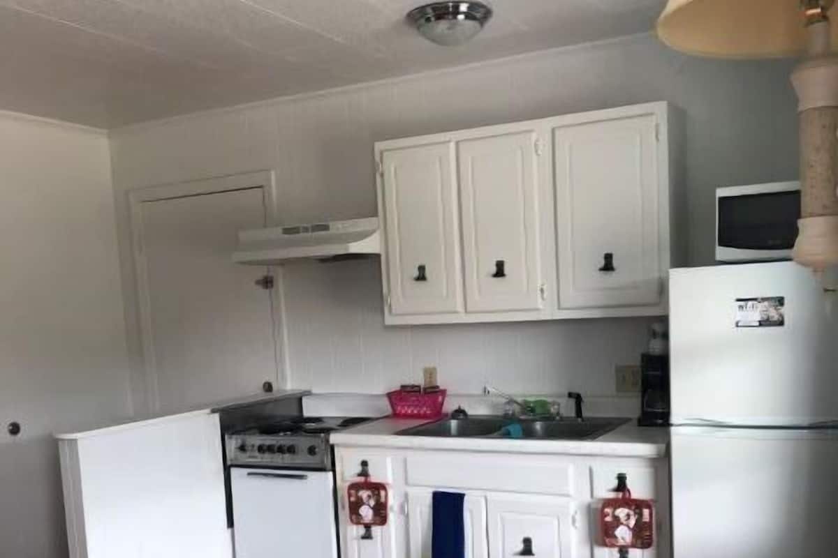 A compact kitchen with white cabinets, a stove, a microwave, and a refrigerator. There is a wooden dining table with chairs.