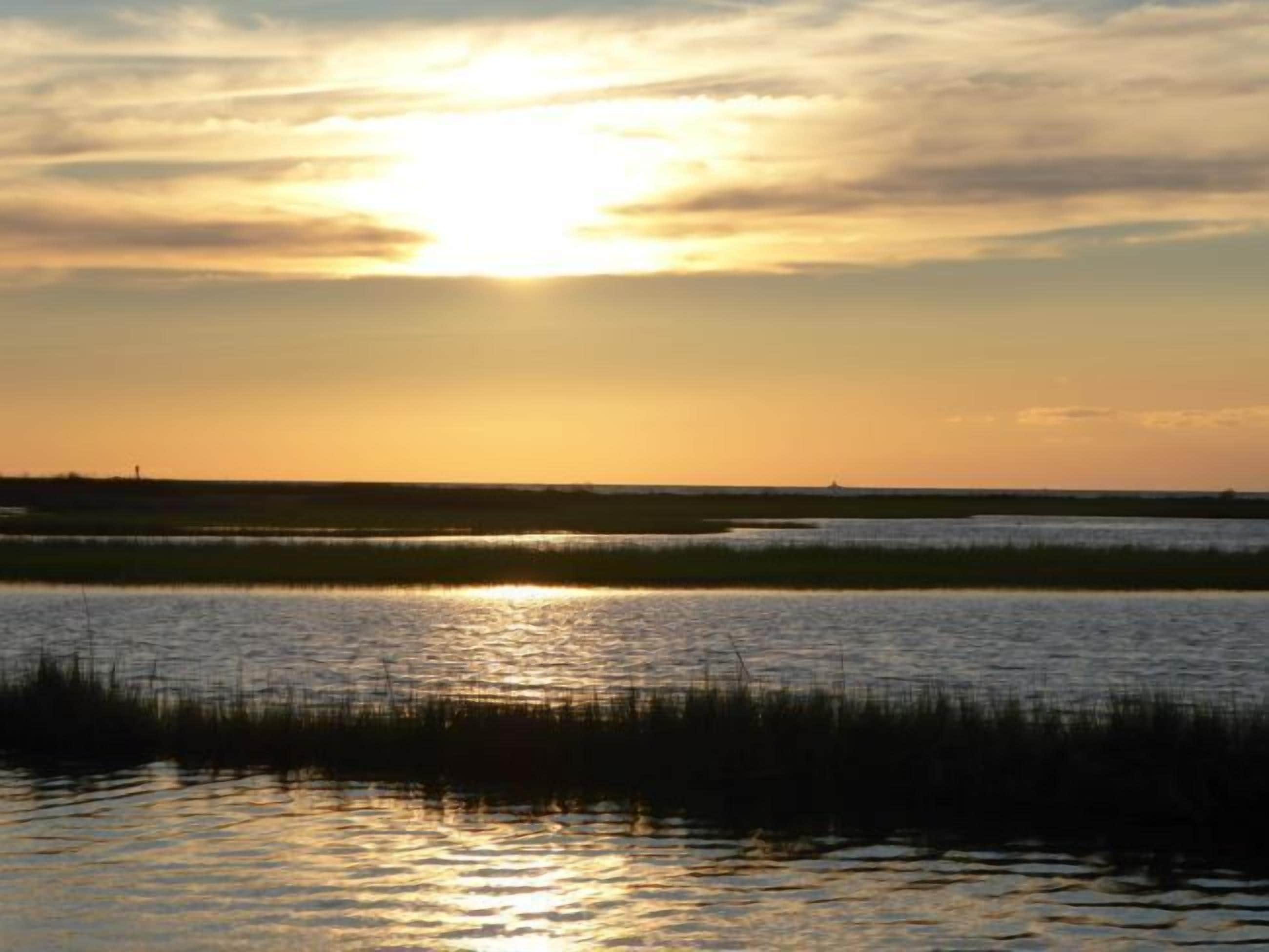 Un coucher de soleil sur une étendue d’eau calme, avec un horizon plat et des bâtiments éloignés.
