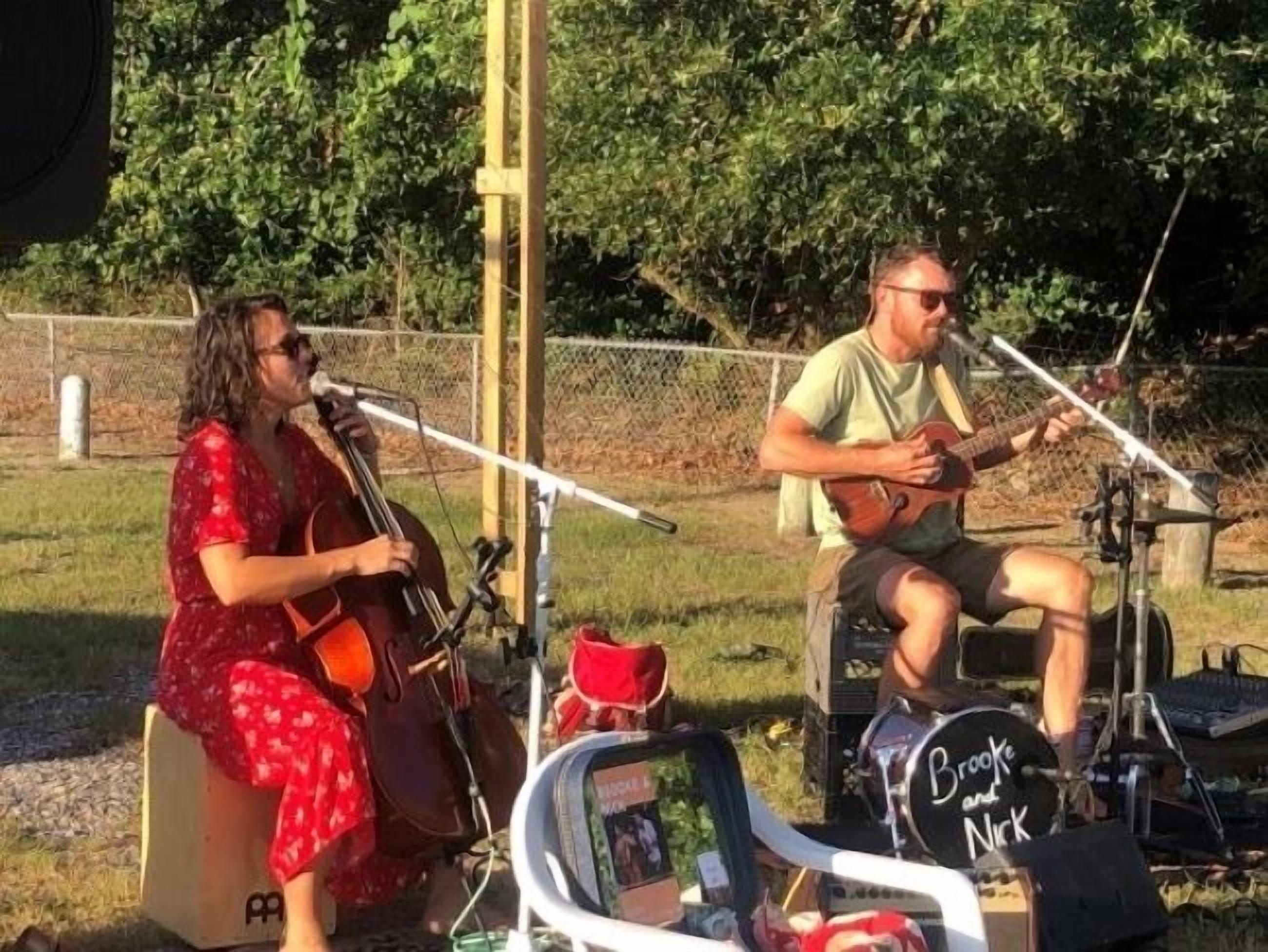 Two people playing musical instruments outdoors, with a fence and greenery in the background.