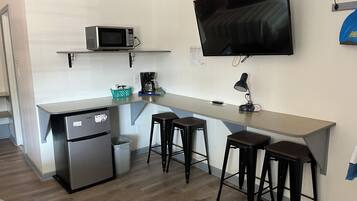 A compact kitchenette with a microwave, coffee maker, and refrigerator. There are three stools and a TV mounted on the wall.