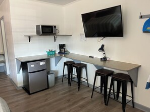 Una kitchenette compacta equipada con microondas, cafetera y refrigerador. Hay tres taburetes y un televisor montado en la pared.