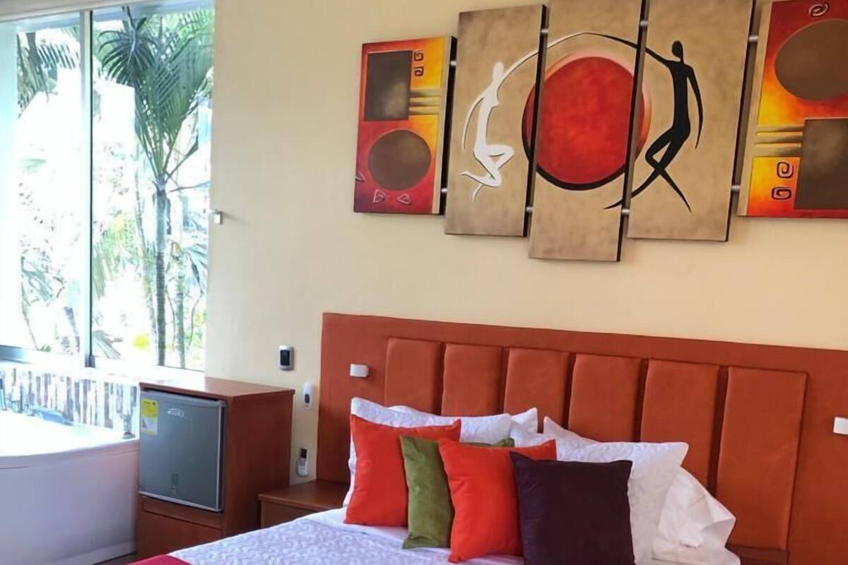 A hotel room with a large bed, a red blanket, and a view of palm trees through the window.