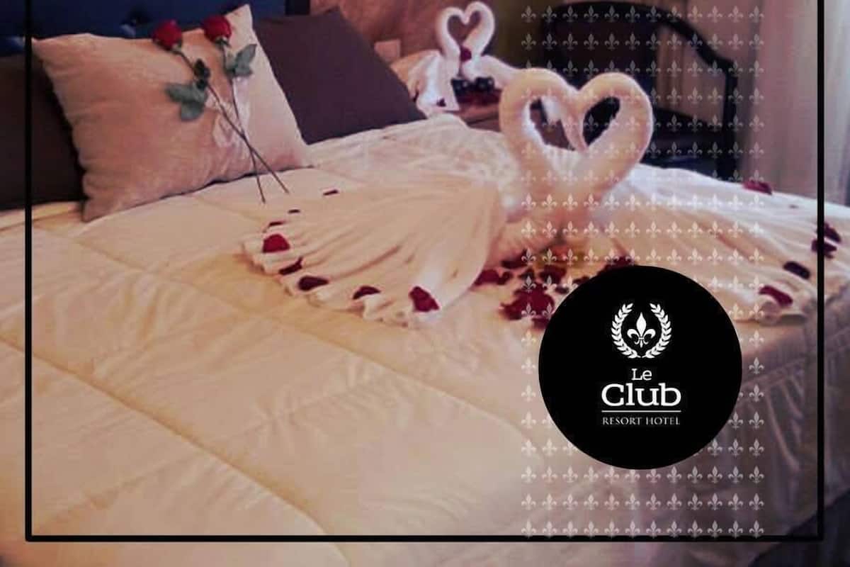 A hotel room with a neatly made bed, a tufted headboard, and a decorative heart-shaped arrangement of towels and flowers.