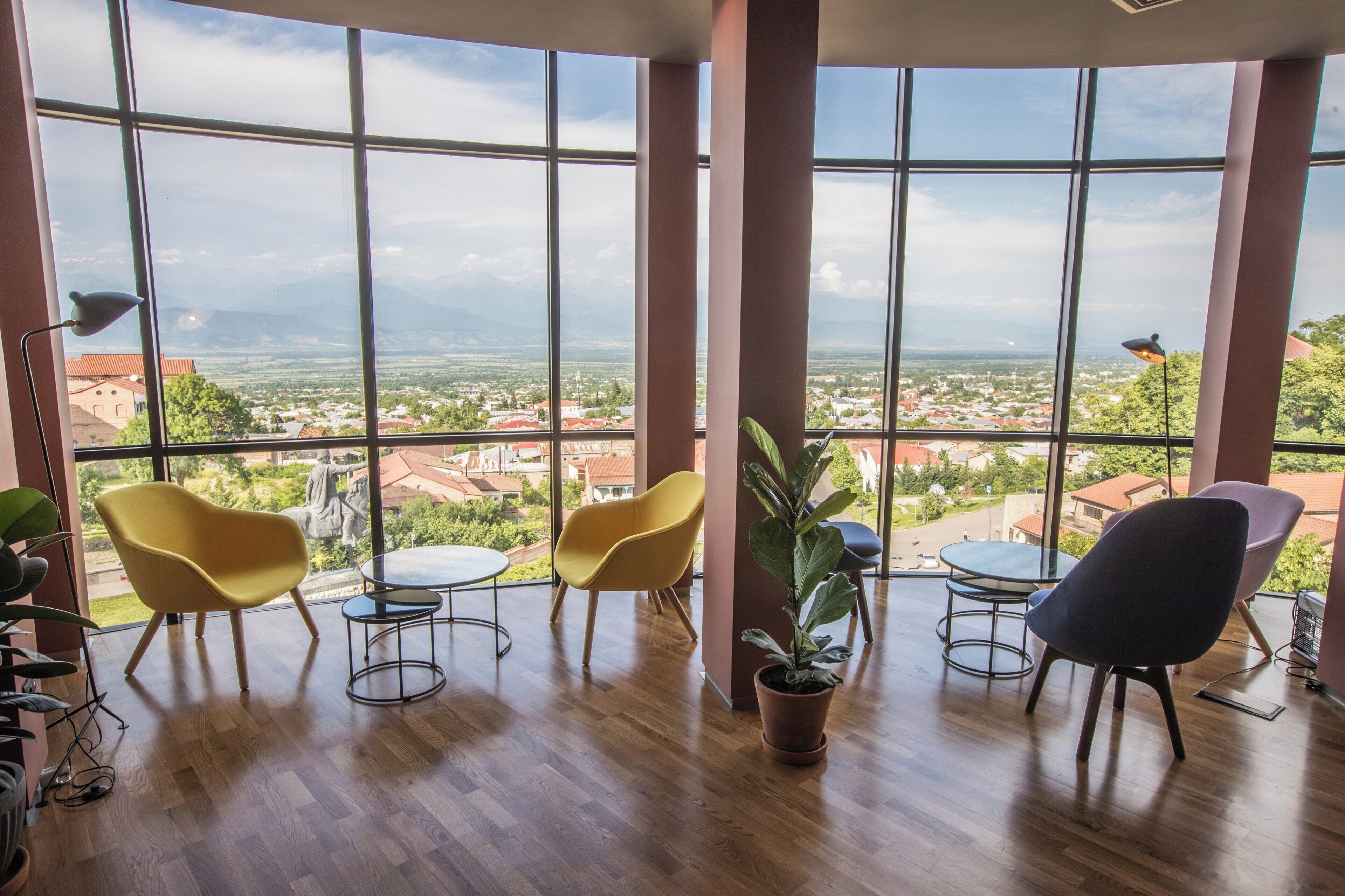 Amplia habitación con ventanas grandes que ofrecen vistas a un paisaje urbano y montañas. Hay cuatro sillas y una mesita, con una planta en maceta en el centro.