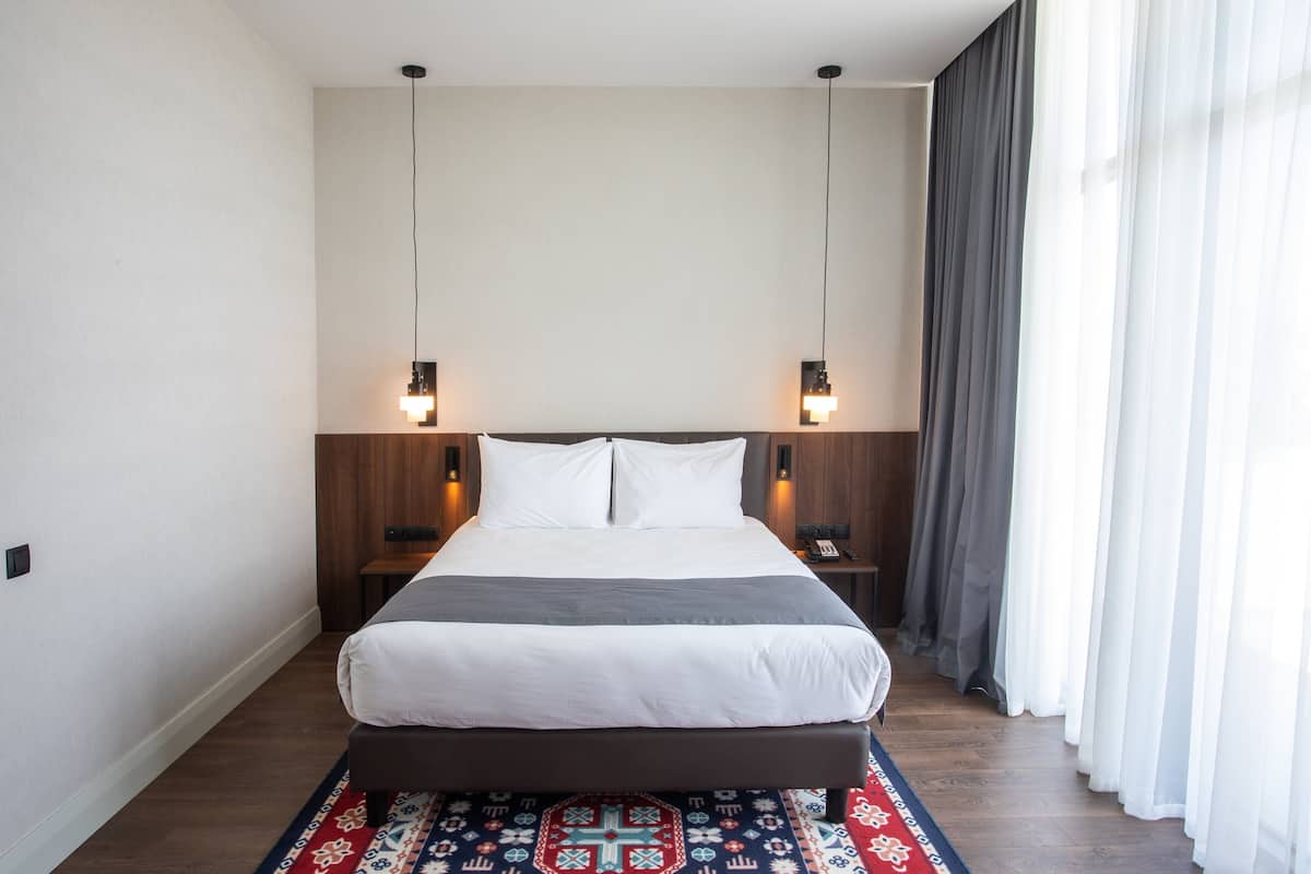 Un lit bien fait, avec une couette blanche et grise, deux tables de chevet avec des lampes et un tapis à motifs sur un plancher en bois.
