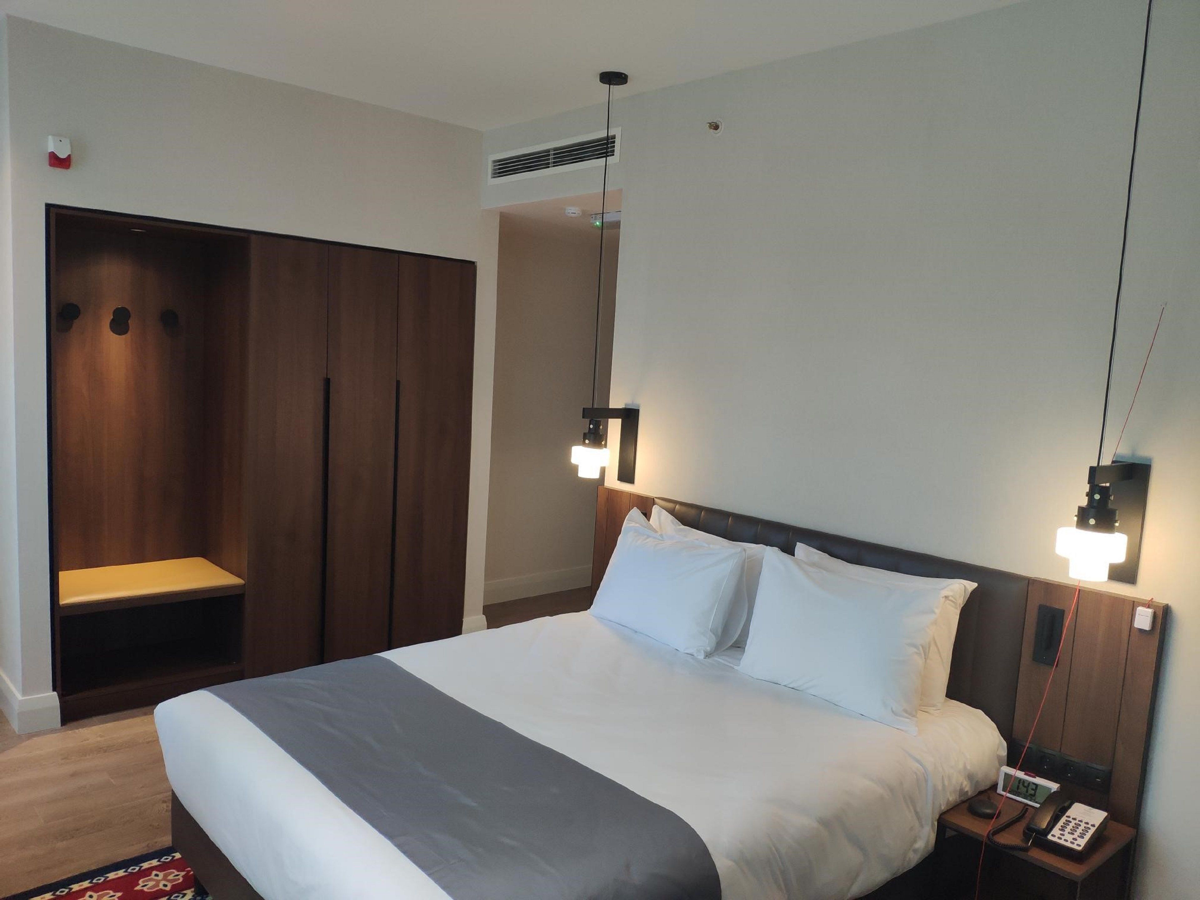 Hotellihuoneessa on suuri sänky, yöpöytä ja vaatekaappi. Sängyn yläpuolella on kaksi seinään kiinnitettyä valaisinta, ja huoneessa on myös pieni pöytä, jossa on puhelin ja kello.