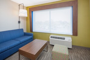 A room with a blue sofa, a wooden table, a patterned ottoman, a standing lamp, and a large window with a view of a cityscape. - Holiday Inn Express & Suites Chico by IHG (Chico)