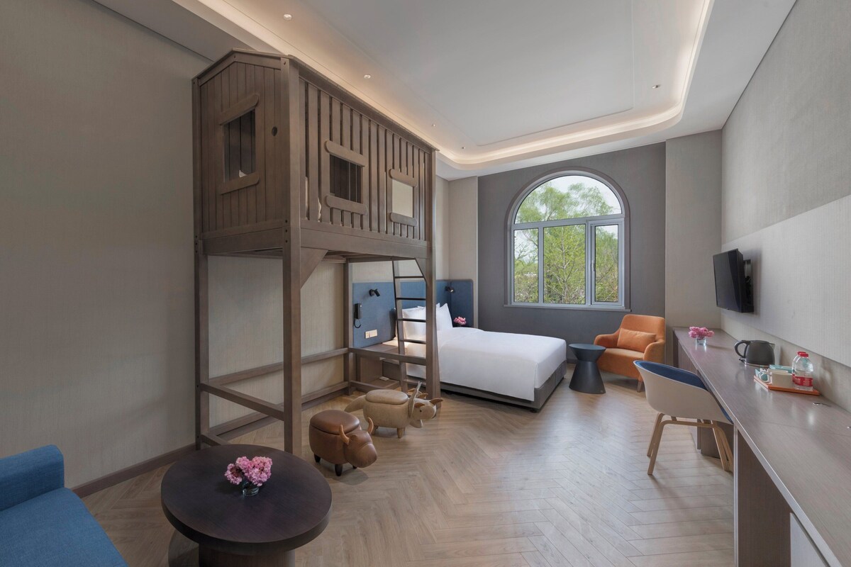 A modern hotel room with a bunk bed, a desk, a chair, a round table with flowers, and a large window with a view of trees.