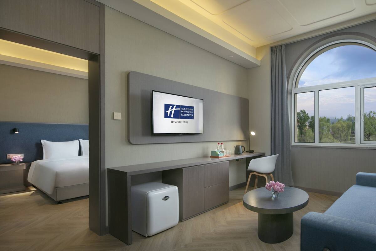 A modern hotel room with a bed, a desk, a small refrigerator, a round coffee table, and a large window offering a view of trees.