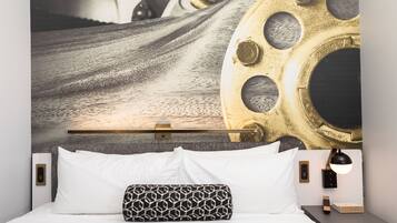 A modern hotel room with a large wall mural, a neatly made bed with white and patterned pillows, a bedside table with a lamp, and a power outlet.