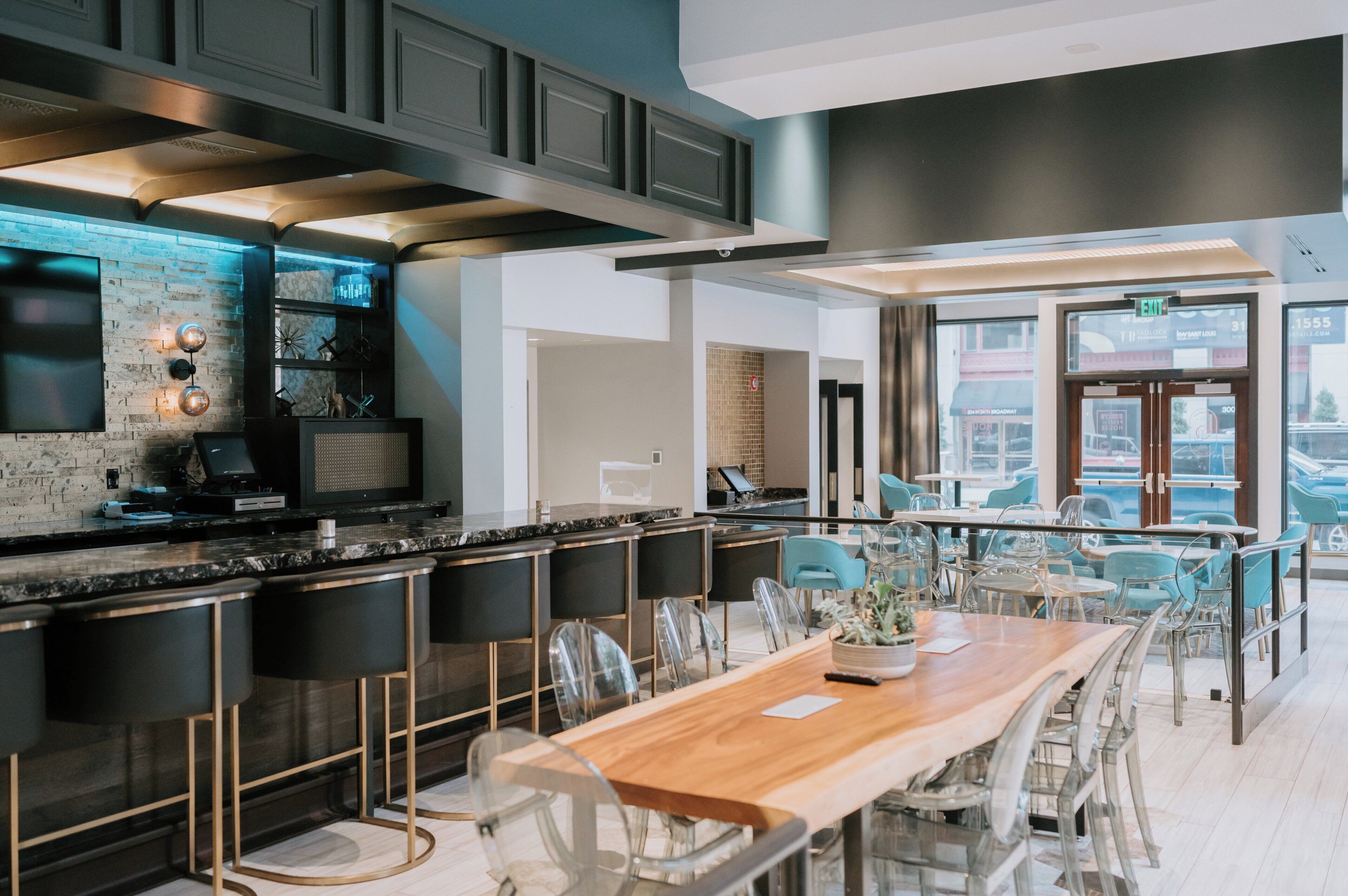 A modern bar area with a long wooden table and clear chairs, a kitchen with a built-in TV and decorative wall lights, and a view of the street through large windows.