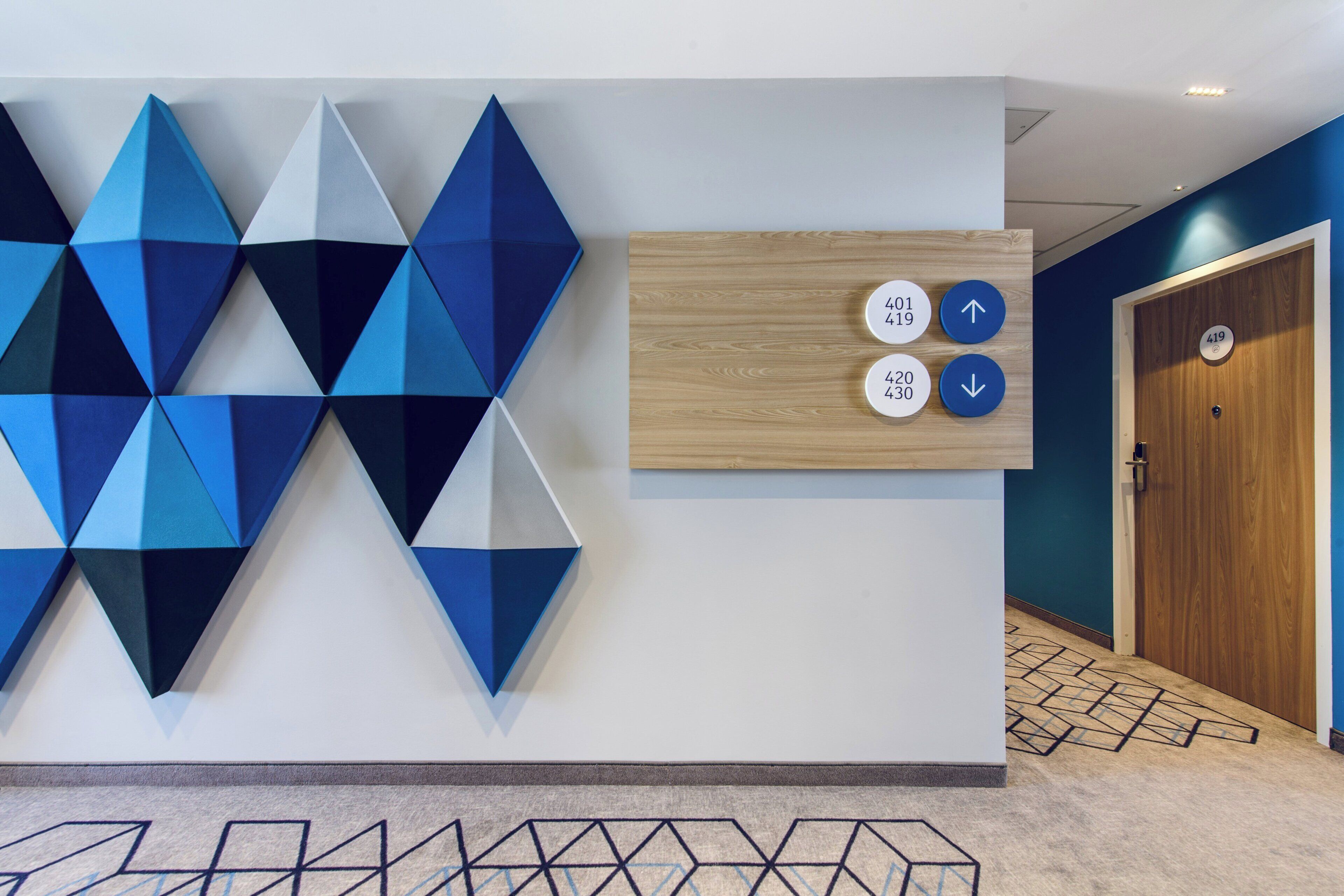 A modern interior with geometric wall art, a wooden panel with room numbers, and a door with a number plate.
