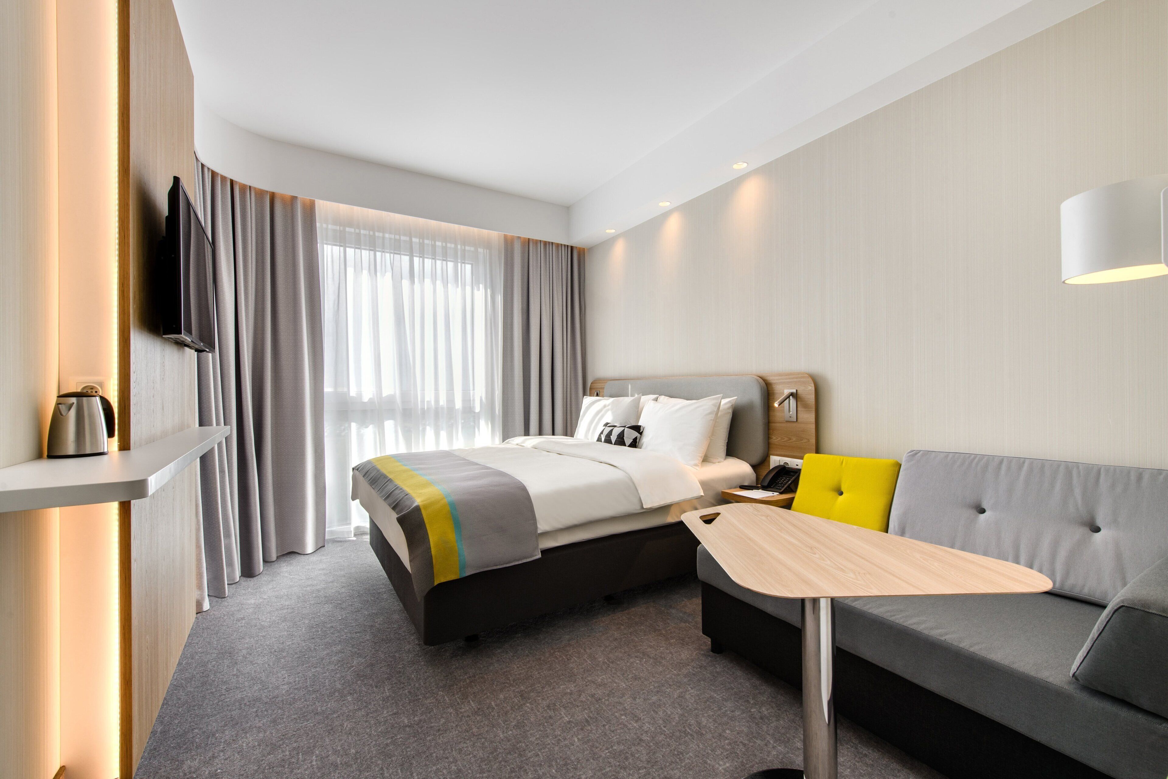 A hotel room with a bed, a sofa, a small table, a TV mounted on the wall, and a window with curtains.