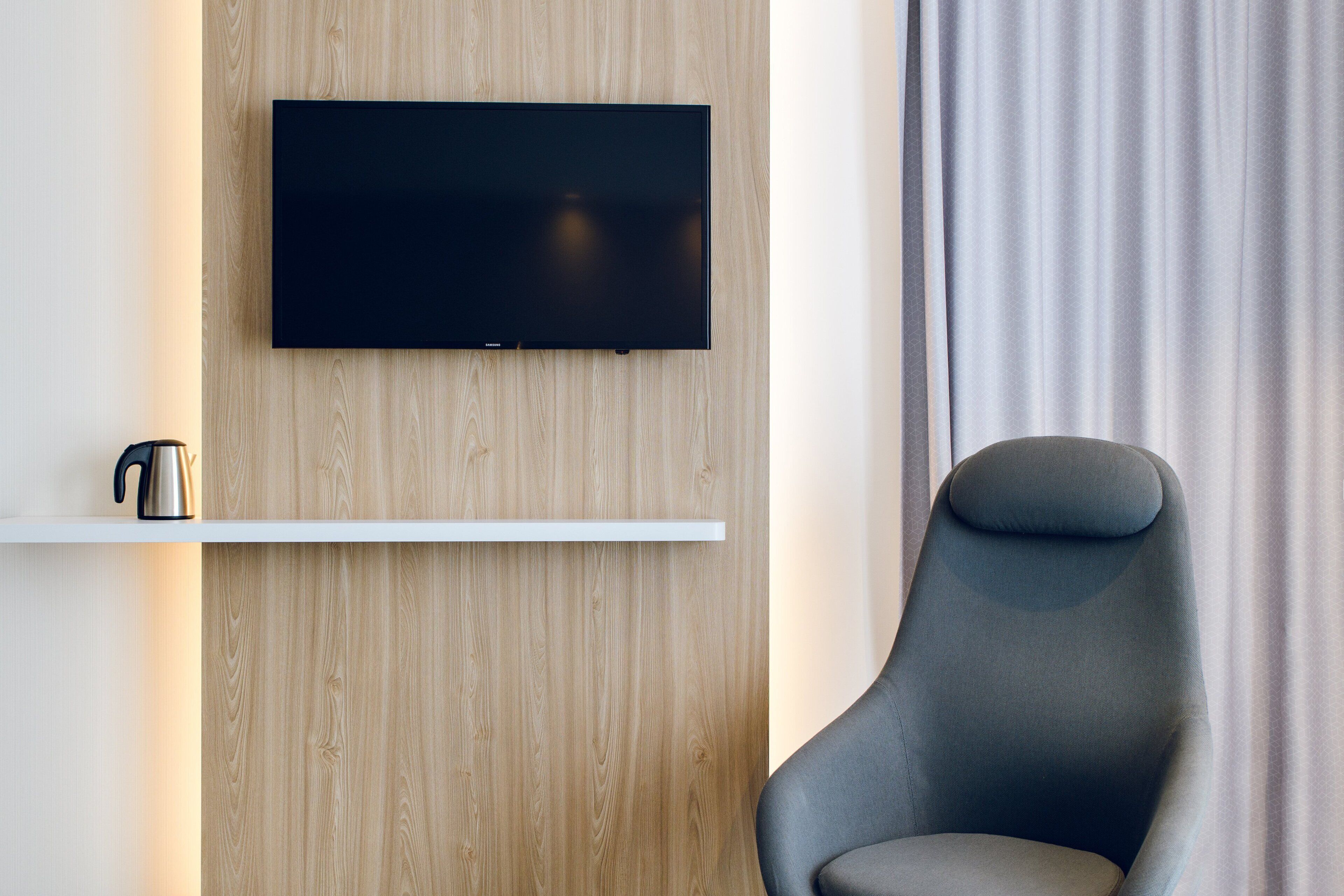 A modern room with a mounted flat-screen TV, a white shelf with a kettle, and a grey armchair.