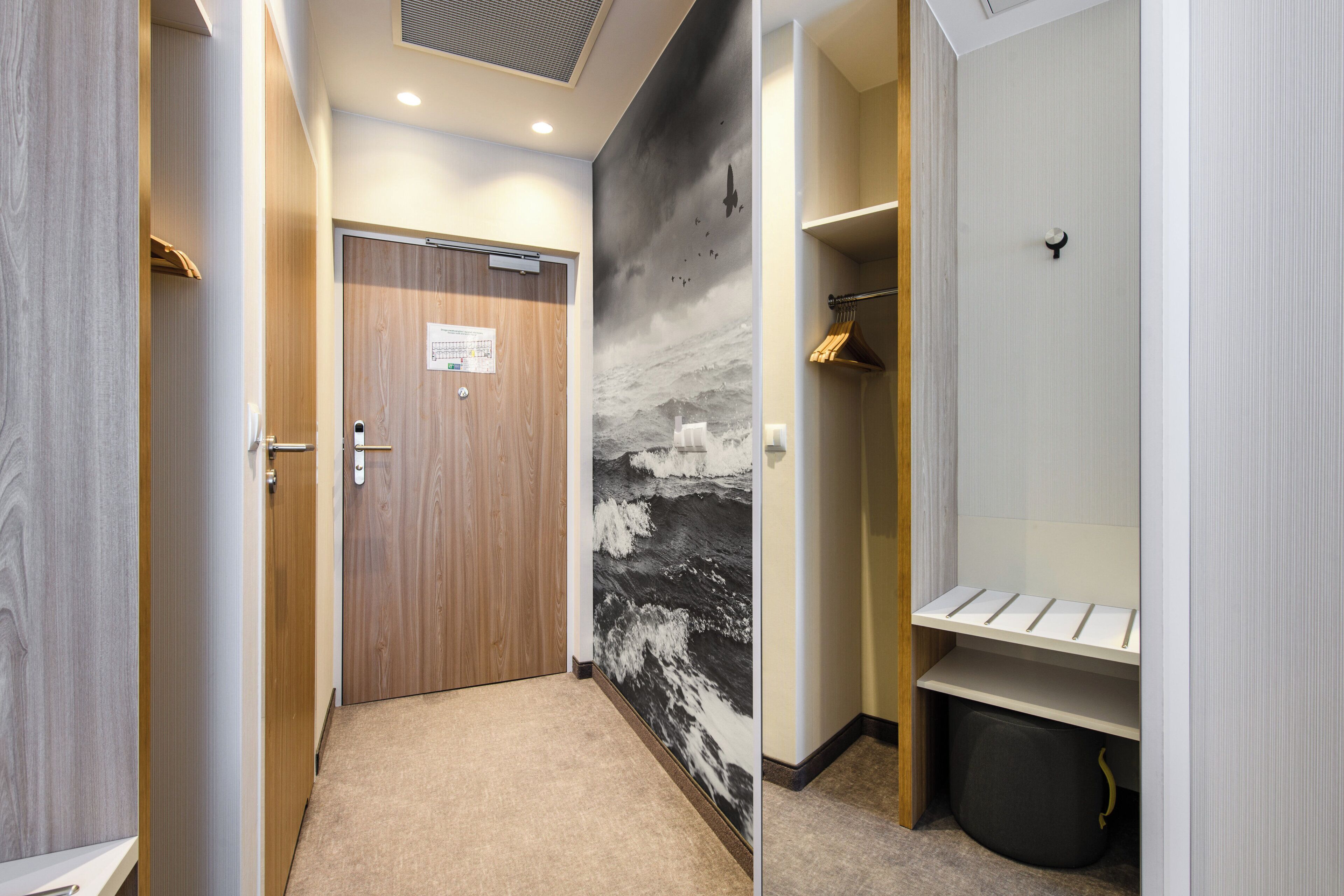 A hallway with a wooden door, a wall with a black and white mural, and a white shelf with a hanging light fixture.