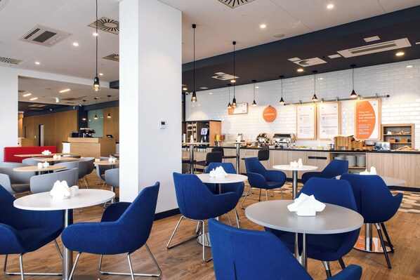 Um café moderno com mesas redondas e cadeiras azuis, uma área de cozinha e um espaço de estar vermelho.