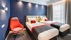 Une chambre d’hôtel avec deux lits, un fauteuil rouge et une grande fenêtre avec des rideaux.