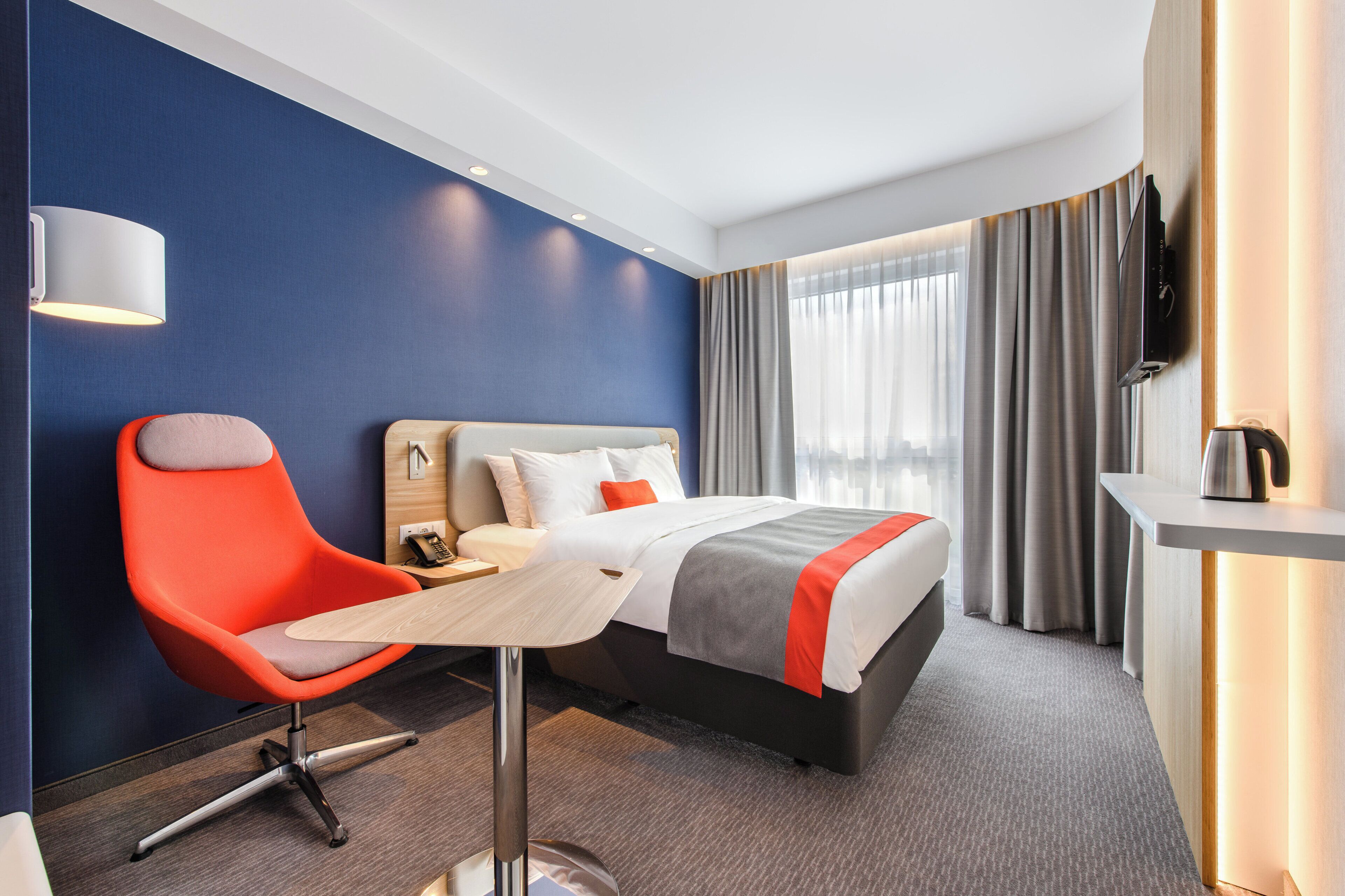 Ein Hotelzimmer mit Bett, Schreibtisch, Stuhl, Fernseher und einem Fenster mit Vorhängen.