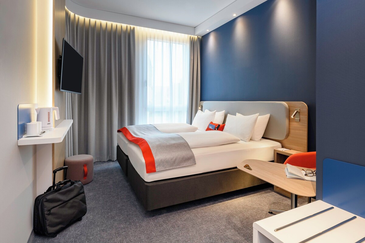 A modern hotel room with a bed, bedside table, TV, and a desk with a chair.