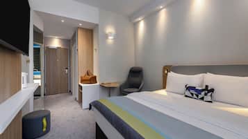 A modern hotel room with a bed, a desk, a chair, a small table, and a wall-mounted TV.
