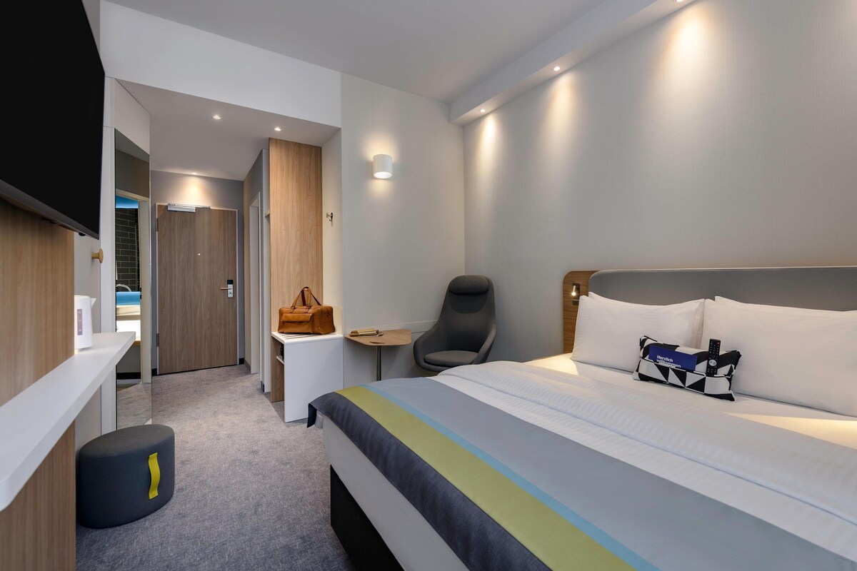 A modern hotel room with a large bed, a desk, a chair, and a small table.