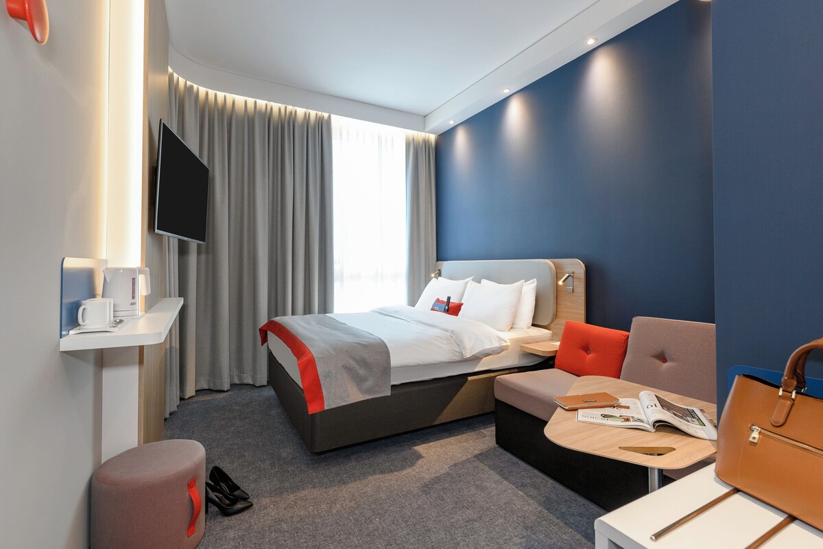A modern hotel room with a bed, seating area, and a flat-screen TV.