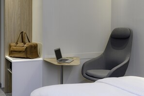 Une chambre d’hôtel moderne avec un lit, un fauteuil gris, une petite table avec un ordinateur portable et un sac à main beige posé sur une commode blanche.