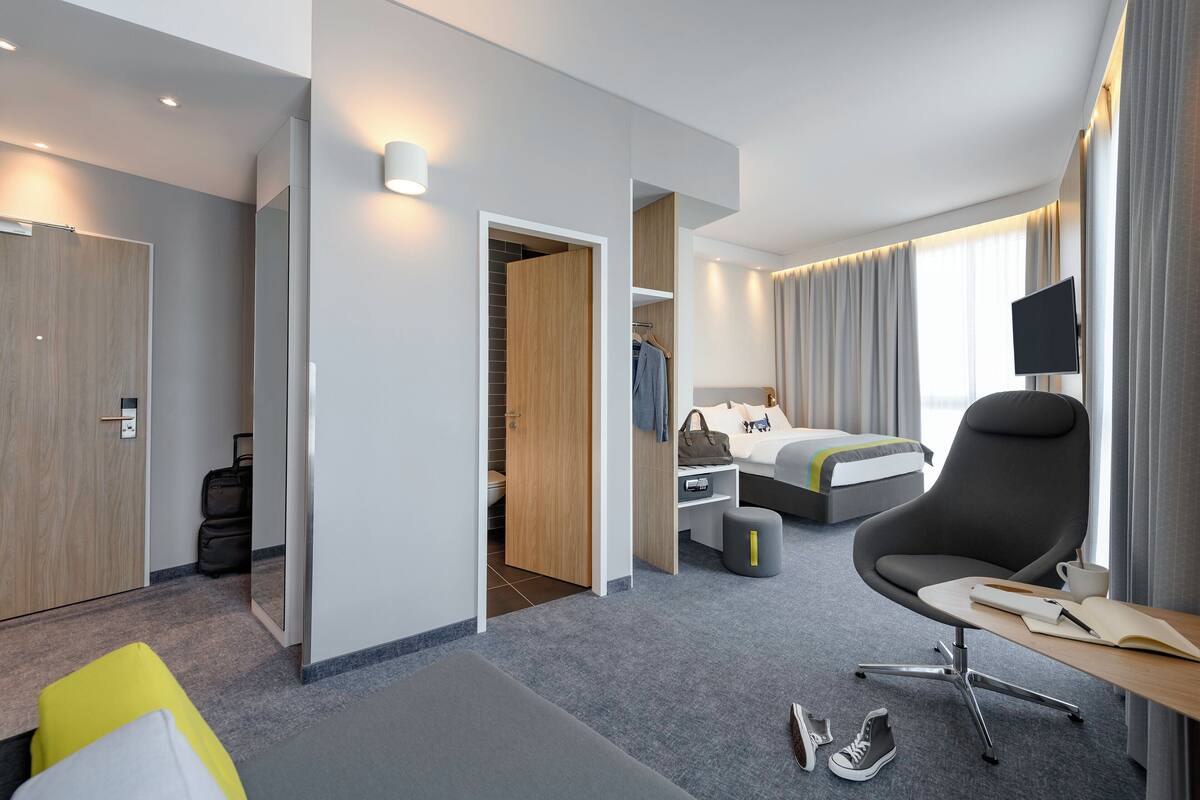 A modern hotel room with a bed, a desk, a chair, a TV, and a wardrobe.
