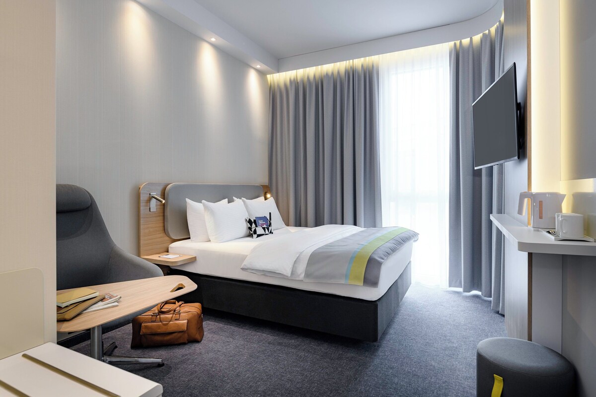 A modern hotel room with a bed, a desk, a chair, a TV, and a window with curtains.