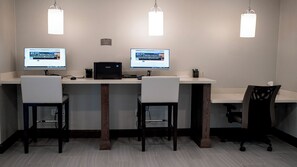 Ein moderner Büroraum mit zwei Schreibtischen, jeweils ausgestattet mit einem Computerbildschirm, einem Drucker und einem Stuhl. Über den Schreibtischen befinden sich zwei Pendelleuchten, und an der Wand hängt ein kleines Schild.
