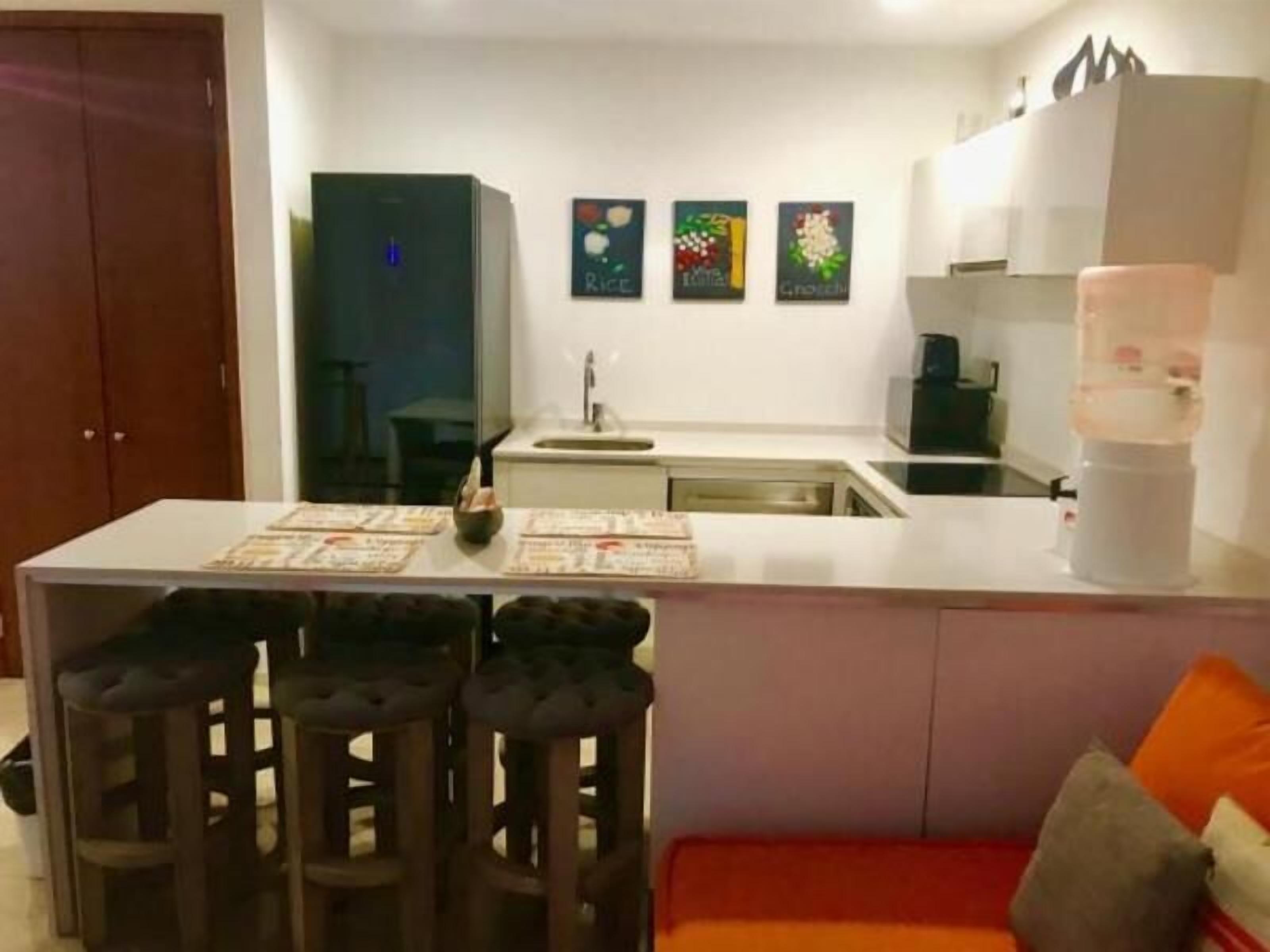 A compact kitchen with a central island, bar stools, a refrigerator, and a coffee machine.