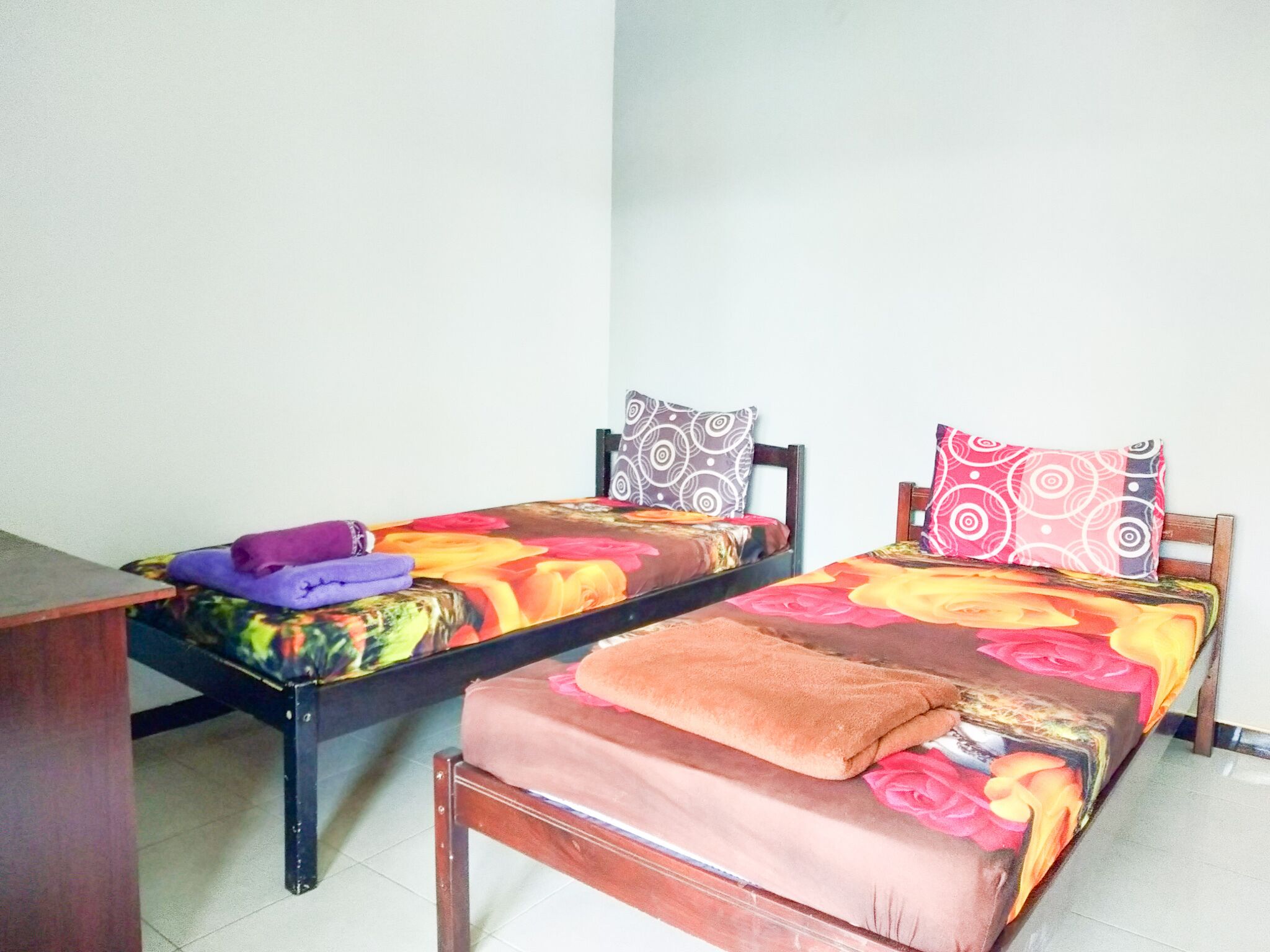 A room with two single beds, each with a patterned bedspread and pillows. There is a wooden table in the foreground.