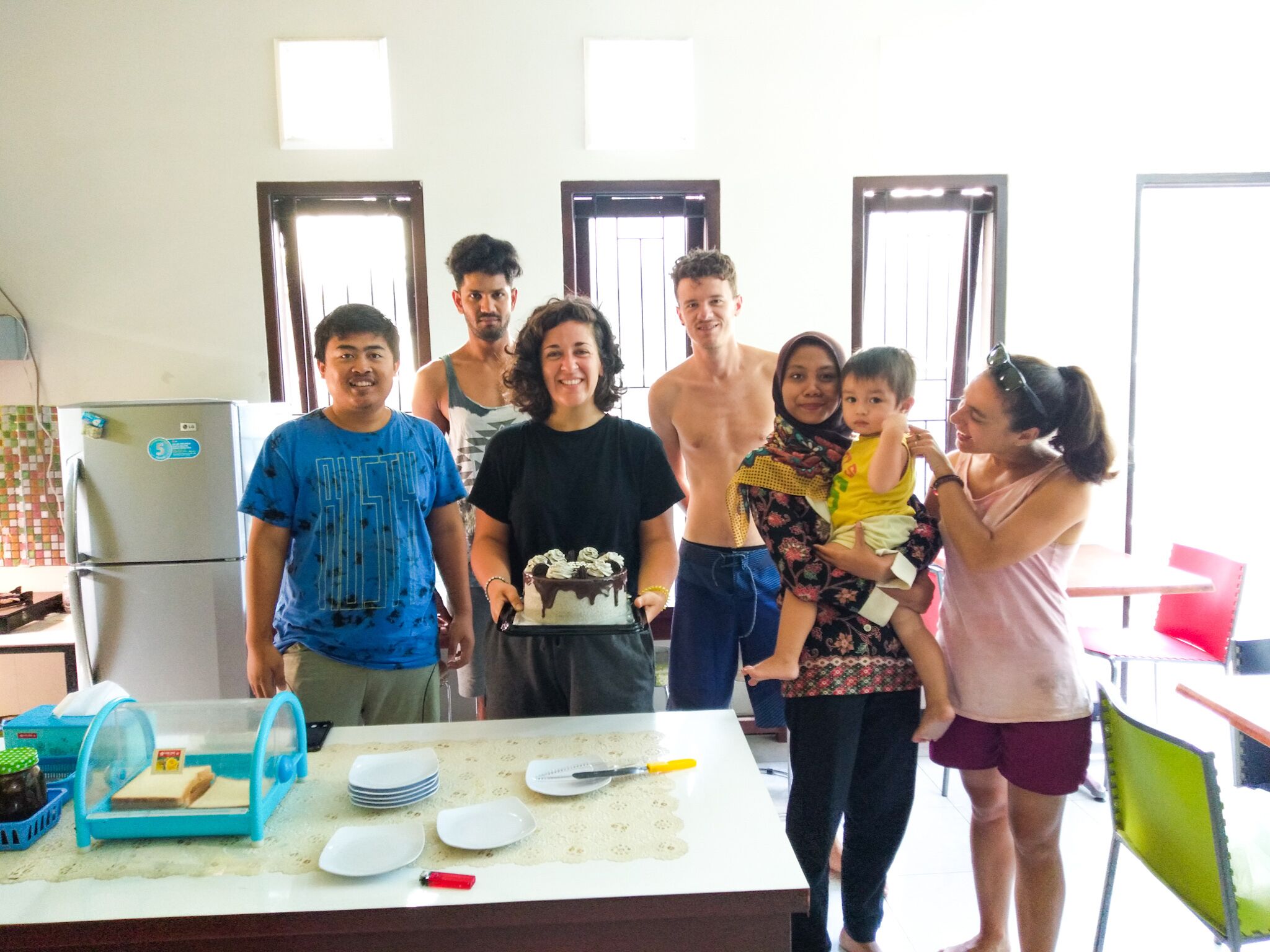 A group of people in a kitchen with a refrigerator, a table with plates and utensils, and a window with blinds.