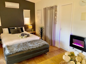 Un dormitorio con cama, lámpara de noche, aire acondicionado, cortinas, un reloj de pared y televisor.