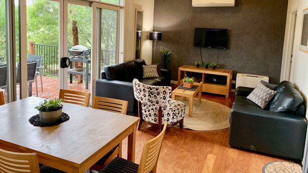 A modern living room with a sofa, coffee table, and TV. There is a balcony with outdoor furniture and a view of trees. - Phillip Island Nature Resort (Cowes)