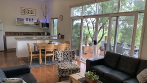 A modern living room with a black leather sofa, a wooden coffee table, and a patterned ottoman. A kitchen with white cabinets and a dining area with wooden chairs is visible. Large windows offer a view of a deck and trees. - Phillip Island Nature Resort (Cowes)