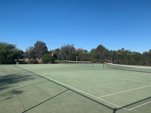 Cancha de tenis al aire libre con una red, rodeada de árboles y un cielo despejado.