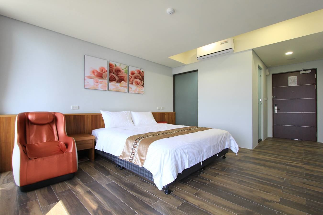 A hotel room with a bed, a red chair, a wooden headboard, a brown patterned throw, and a wall-mounted air conditioner.