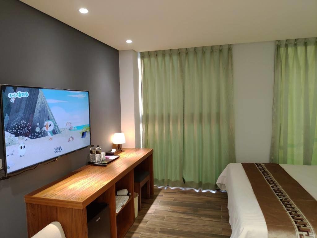 Ein Hotelzimmer mit einem Bett, einem Wandfernseher, einem Holztisch und einem Fenster mit grünen Vorhängen.