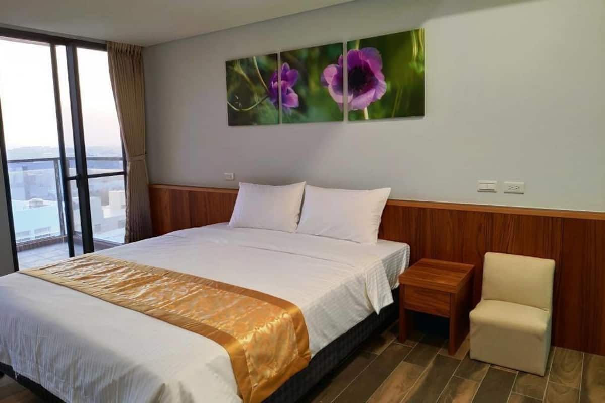 A hotel room with a large bed, a view through a window, a wooden headboard, a bedside table, and a chair.